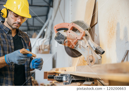 Carpenters man in a yellow hardhat, safety glasses measuring wood for cutting, with a circular saw in the background. Ideal for woodworking and carpentry workshop settings, National Carpenters Day 120200663