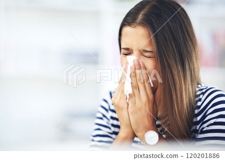 Its allergy season again. Shot of a young woman with allergies sneezing into a tissue at home. 120201886