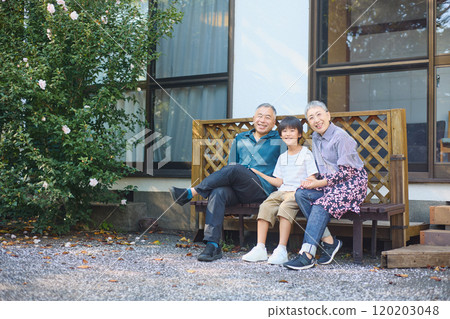 An elderly couple and their grandchild sitting on a bench outside their house 120203048