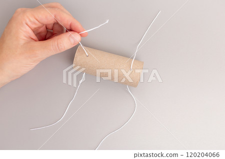 hand is holding toilet paper cardboard roll tube while the other hand is threading a string through holes, part of DIY project, 120204066