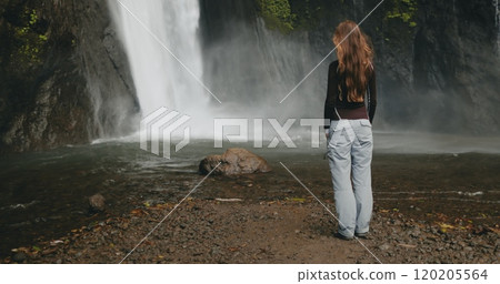 Tourist woman looking powerful Bali waterfall, water cascading down rocks. Long hair blows in wind. Tourism, trekking, hiking, relax, active lifestyle, summer holiday vacation. Slow back view Tourist woman looking powerful Bali waterfall, water cascading down rocks. Long hair blows in wind. Tourism, trekking, hiking, relax, active lifestyle, summer holiday vacation. Slow back view 120205564