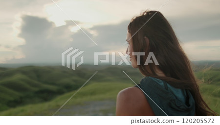 Young woman stands on a hill in indonesia, watching the sunset over lush green hills, painting the sky with orange and pink hues 120205572