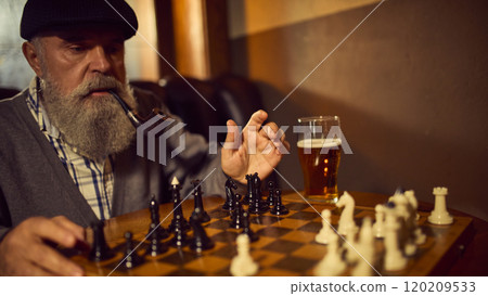 Bearded elderly man in a cap, smoking tobacco pipe and playing chess with his friend, drinking beer in cozy vintage bar 120209533