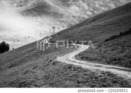 A winding path ascends through lush alpine grass, guiding hikers toward a mountain trail. A ski lift gently hovers above, suggesting an inviting summer adventure amid nature. 120209851