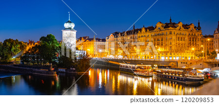 The Sitkov Water Tower stands tall along Masaryk Embankment in Prague, Czechia, as the sun sets and the city lights begin to illuminate the scene. The calm water reflects warm glow of the buildings. 120209853