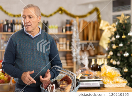 Mature male shopper with grocery basket attentively choosing fresh food and alcohol in grocery supermarket for Christmas celebration 120210395