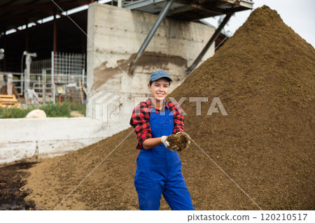 Girl worker in blue jumpsuit stands next to pile of manure. 120210517