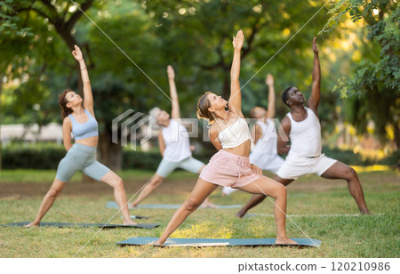 Girl performing Baddha Viparita Virabhadrasana during group yoga in park 120210986