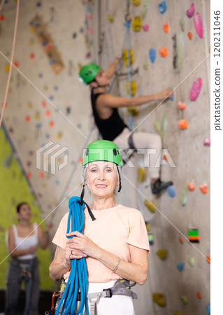 Smiling active mature woman standing with rope on shoulders and helmet in hand against artificial training climbing wall in adventure park Smiling active mature woman standing with rope on shoulders and helmet in hand against artificial training climbing wall in adventure park 120211046