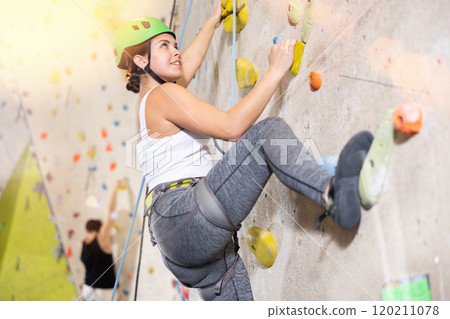 Confident young woman wearing helmet scaling artificial climbing wall in climbing gym Confident young woman wearing helmet scaling artificial climbing wall in climbing gym 120211078