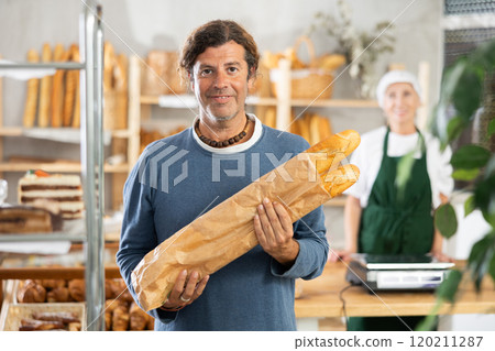 Man buyer with baguettes in package stands near showcase in bakery Man buyer with baguettes in package stands near showcase in bakery 120211287