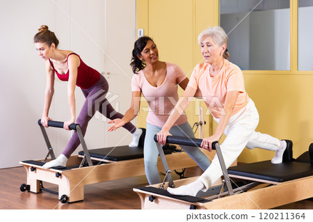 Aged woman doing pilates on reformer with Hispanic female trainer Aged woman doing pilates on reformer with Hispanic female trainer 120211364