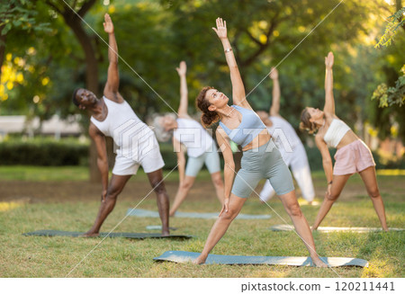 Multinational yoga enthusiasts with female coach perform Trikonasana in park 120211441