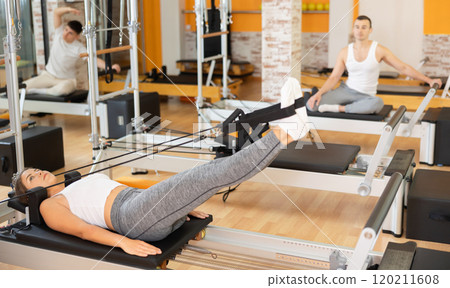 Young woman doing Pilates on reformer with straps on feet Young woman doing Pilates on reformer with straps on feet 120211608