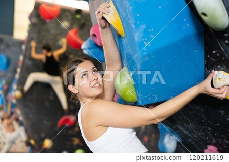 Positive focused young woman exercising on black climbing wall without safety belts and preparing for summer mountain hiking in game center indoors 120211619