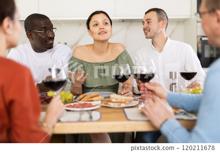 Middle-aged friends sitting at table with a glass of wine while having friendly conversation Middle-aged friends sitting at table with a glass of wine while having friendly conversation 120211678