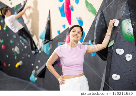 Young smiling Asian girl coach stands at climbing wall and leans holding on to hook. Young smiling Asian girl coach stands at climbing wall and leans holding on to hook. 120211680