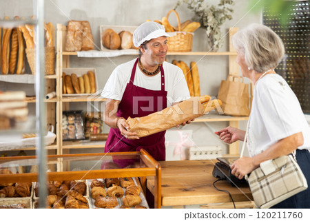 Man entrepreneur, owner of baker puts baguettes in paper bag, serves senior female client 120211760