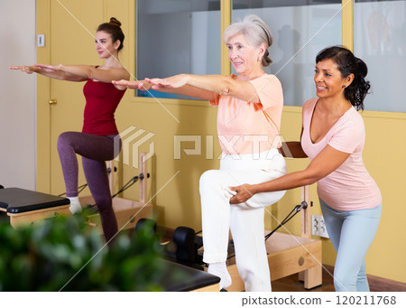 Hispanic woman pilates instructor helping aged woman exercising on wunda chair 120211768