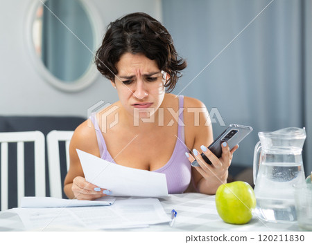 Shocked young woman looking at document and holding smartphone in hand Shocked young woman looking at document and holding smartphone in hand 120211830