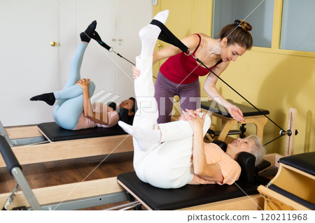 Young pilates instructor assisting elderly woman to do exercises on reformer 120211968