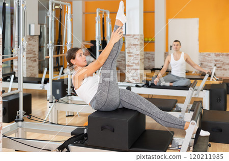 Girl works out in gym and performs set of exercises to stretch leg muscles with help of reformer 120211985