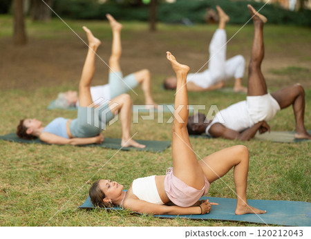 Multinational yoga enthusiasts with female coach perform Setu Badha Sarvangasana in park Multinational yoga enthusiasts with female coach perform Setu Badha Sarvangasana in park 120212043