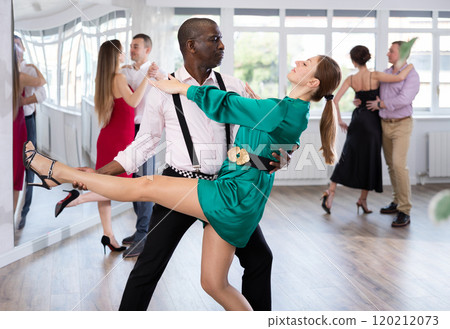 Elegant duet of man and woman in festive dresses dances passionate tango during group dance lesson in choreographic studio 120212073