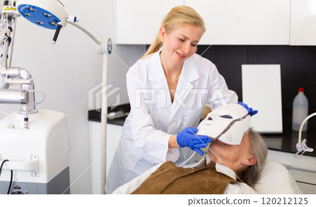 Cosmetologist putting spectrum mask on senior woman's face 120212125