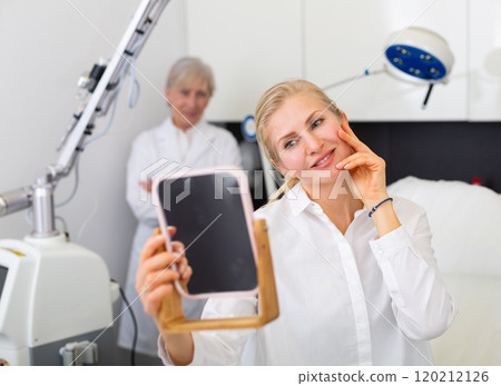 Female client looking in mirror satisfied with results of cosmetology procedure 120212126