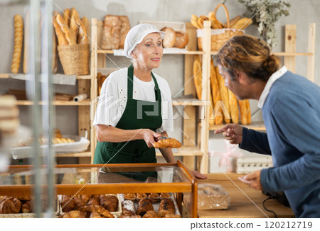 Elderly woman seller offers buyer to choose croissant, shows finished products. 120212719