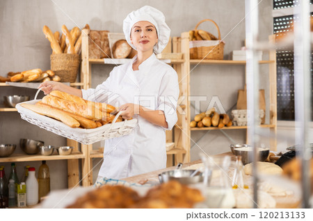 European young bakery girl seller in white uniform and and baker's hat standing proudly in front of fresh bread holding basket of freshly baked baguettes in kitchen 120213133