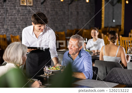 Young waiter serving ordered dishes to senior couple in restaurant Young waiter serving ordered dishes to senior couple in restaurant 120213222