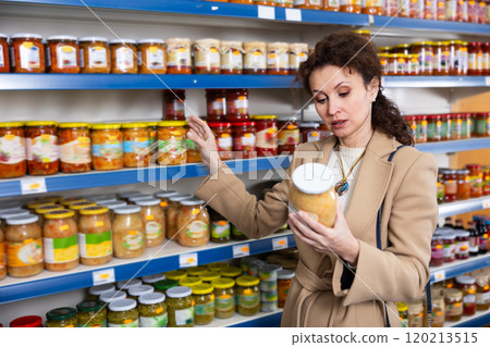 Female shopper selects glass jar of sauerkraut at grocery store. Woman shopper reading expiration date on product label 120213515