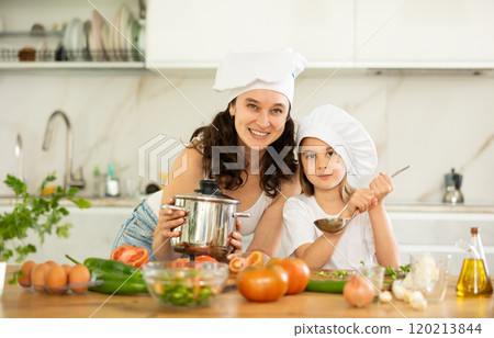 Mother and girl in white cooking hats are standing in kitchen near table with dishes. 120213844