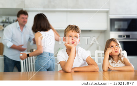 Two children sitting at the kitchen table sadly while their parents quarrelling Two children sitting at the kitchen table sadly while their parents quarrelling 120213977