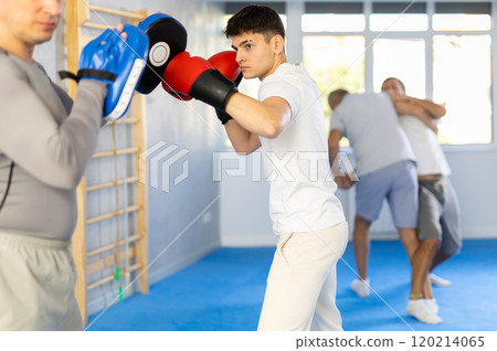 Coach holding mitts during self defence training in gym with young guy mastering base punches in boxing gloves in gym Coach holding mitts during self defence training in gym with young guy mastering base punches in boxing gloves in gym 120214065
