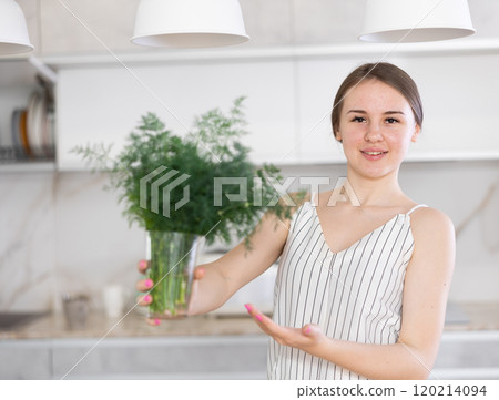 Woman in dress with short sleeves sitting near kitchen table and holding glass with bunch of dill Woman in dress with short sleeves sitting near kitchen table and holding glass with bunch of dill 120214094
