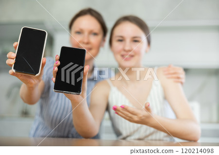 Mother and daughter showing their phones sitting at kitchen-table Mother and daughter showing their phones sitting at kitchen-table 120214098