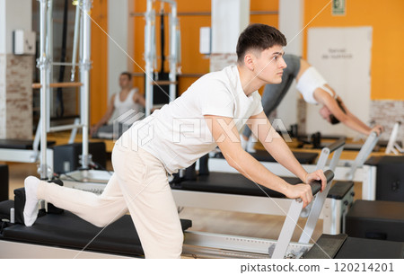 Guy doing stretching exercises on Pilates reformer in fitness center 120214201