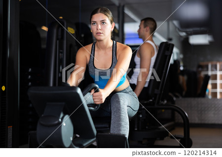 Sportive young girl training her arms using cable exercise machine in gym 120214314