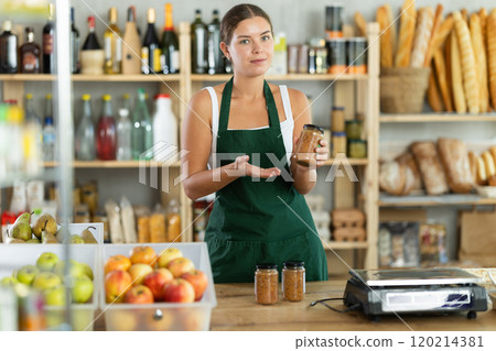 Portrait of friendly girl seller behind a store counter offering to buy canned lentils Portrait of friendly girl seller behind a store counter offering to buy canned lentils 120214381