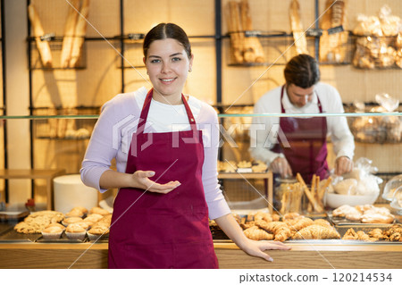 Cheerful young bakeshop salesgirl in maroon apron showing pastries 120214534
