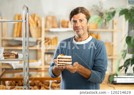 Pleased middle-aged guy holding pastry on paper plate in bakery 120214629