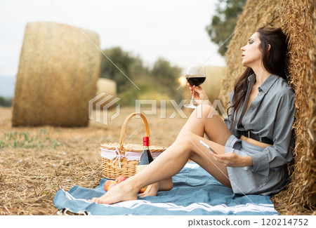 Girl in shirt holds glass of wine and phone sitting on plaid near stack of straw Girl in shirt holds glass of wine and phone sitting on plaid near stack of straw 120214752