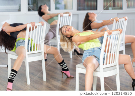Beautiful young woman practicing strip plastic movements while sitting on chair and graceful leg stretch workout with group of athletic women at dance class Beautiful young woman practicing strip plastic movements while sitting on chair and graceful leg stretch workout with group of athletic women at dance class 120214830