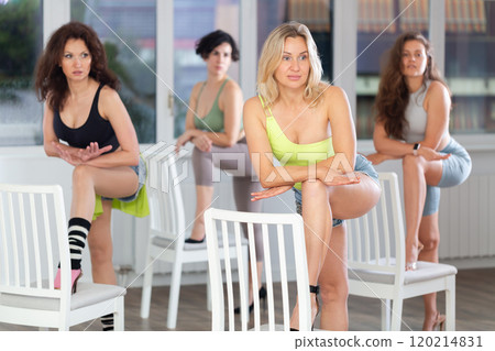 Graceful happy woman in casual activewear confidently leading strip class with dancers following her movements in dance studio, putting leg on chair and demonstrating high heels dance moves 120214831
