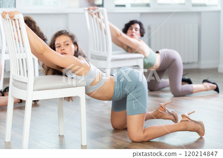 Young elegant female dancers in sportswear and high heels performing kneeling dance moves near chair on floor in choreographic studio during group lesson Young elegant female dancers in sportswear and high heels performing kneeling dance moves near chair on floor in choreographic studio during group lesson 120214847