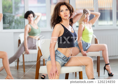 Attractive young woman blending sensuality with fashion while practicing seductive movements sitting on chair in group high heels dance class with sports women at dance studio 120214850
