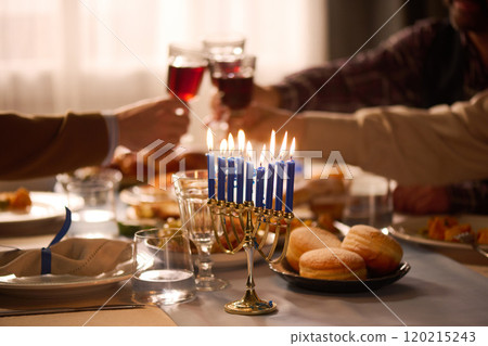 Medium close up of lit blue candles in golden candlestick on festive table, unrecognizable friends clinking glasses with red wine in blurred background 120215243